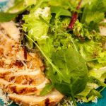 Close-up of a grilled chicken salad with mixed greens and cheese, served on a blue plate.
